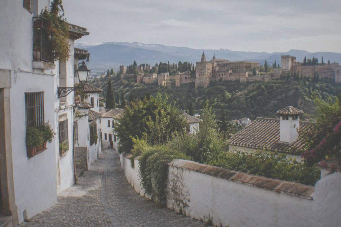 Narrow whitewashed lanes of the Albaicin quarter climbing a hillside in Granada, with the Alhambra visible in the distance.