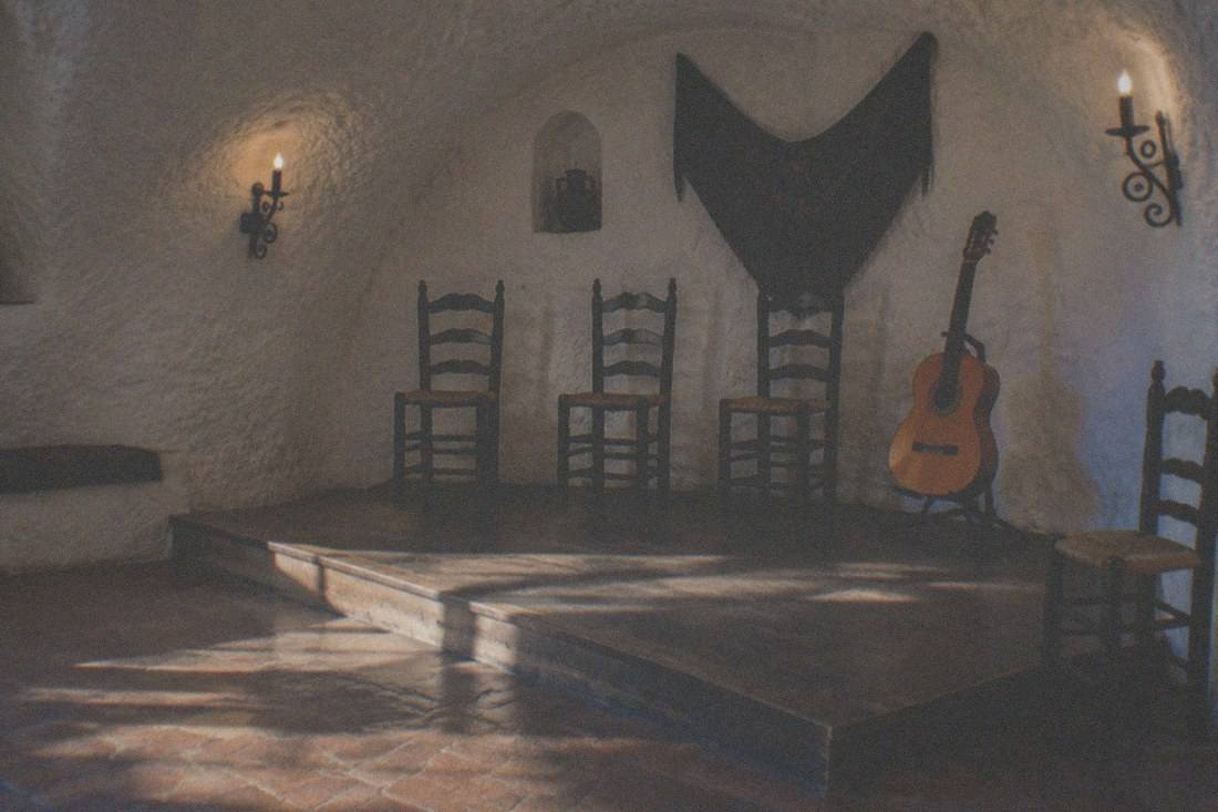 The whitewashed stone interior of a Sacromonte cave venue set for a flamenco performance, with wooden chairs and guitars on display.