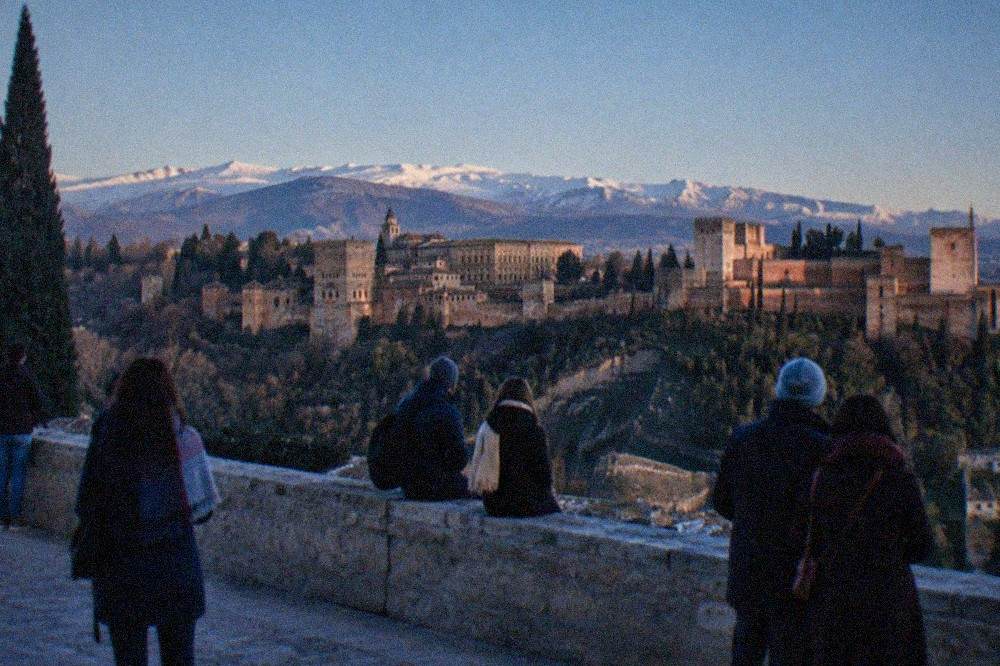 People gathered at the Mirador de San Nicolas at dusk looking across at the Alhambra with snowy Sierra Nevada peaks behind it