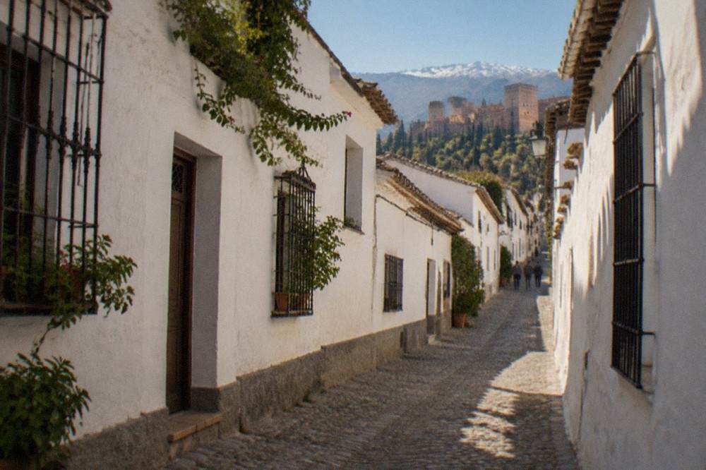 A narrow white-walled street in the Albaicin quarter of Granada with the Sierra Nevada visible in the distance