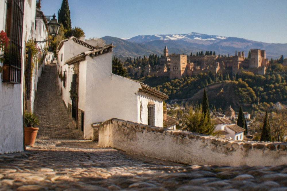 Sunset view from a hilltop square across the Darro valley toward the Alhambra with mountains behind
