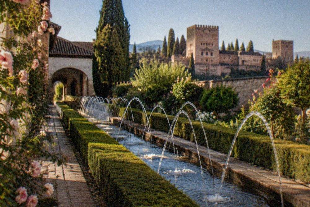 A lit pathway through the Generalife gardens of the Alhambra with hedges and fountains visible in the evening light