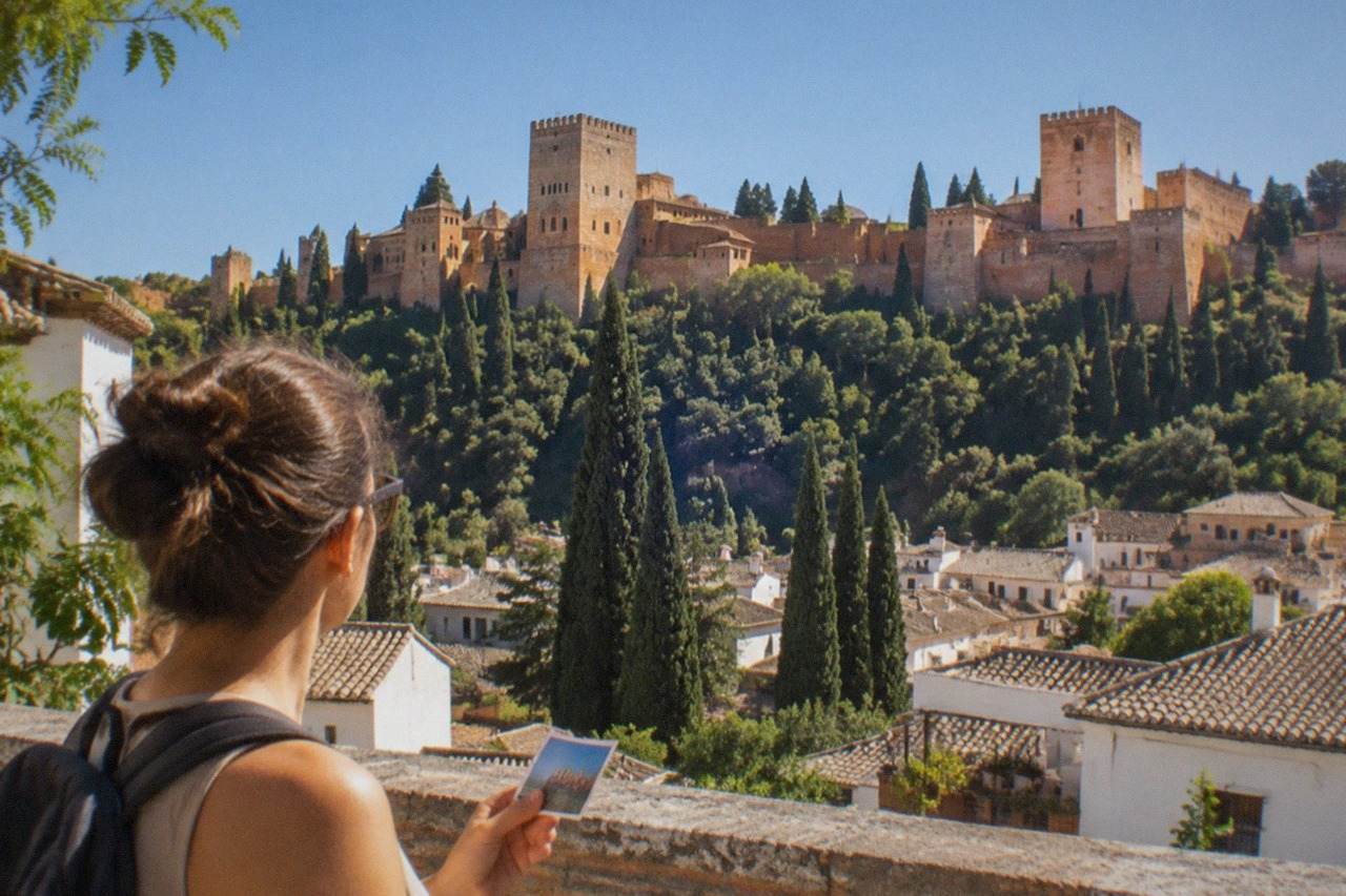 A visitor holding a city pass while looking across Granada's rooftops toward the Alhambra on the hillside.