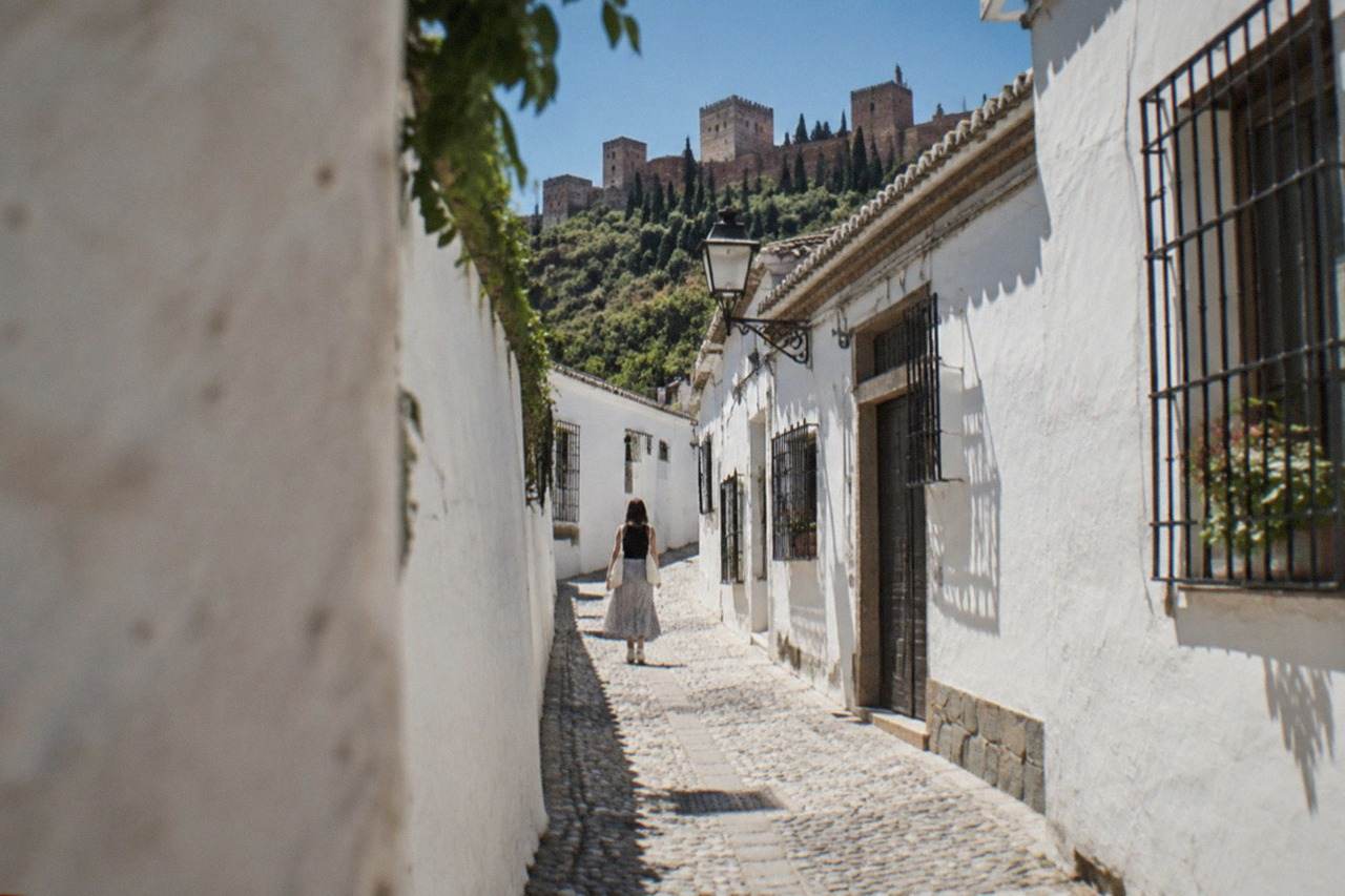 A narrow whitewashed street in the Albaicin quarter of Granada with the Alhambra visible on the hillside above
