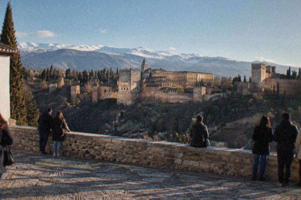 An evening view from the Mirador de San Nicolas with the illuminated Alhambra across the valley and people gathered at the railing