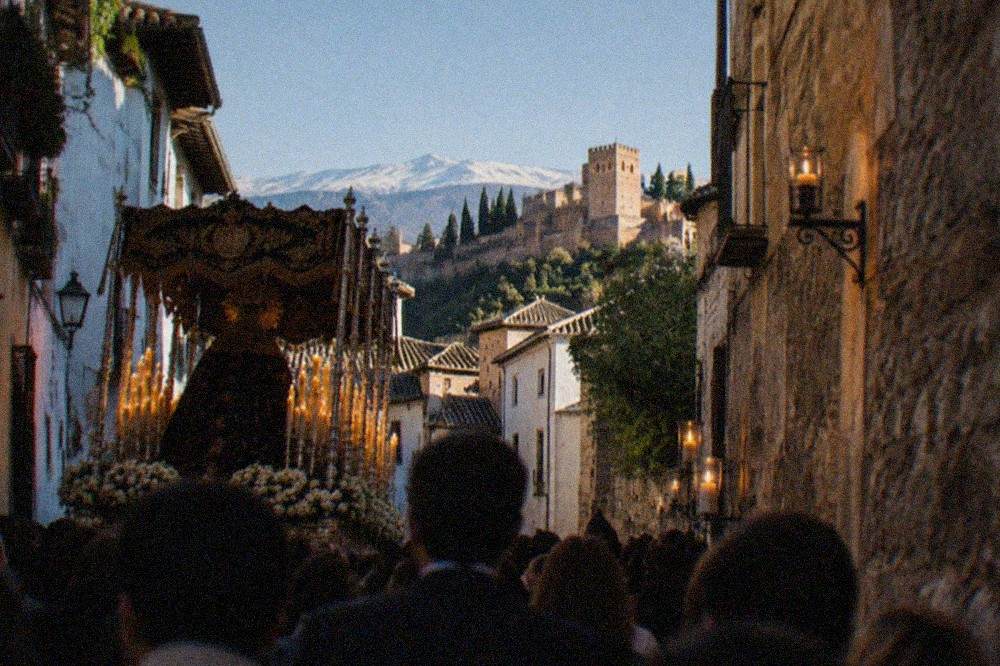 An evening procession crowd in a narrow Granada street, with a decorated float and candlelight on old stone walls