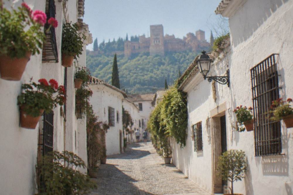 A narrow white-walled lane in the Albaicin quarter with flower pots and the Alhambra visible in the distance