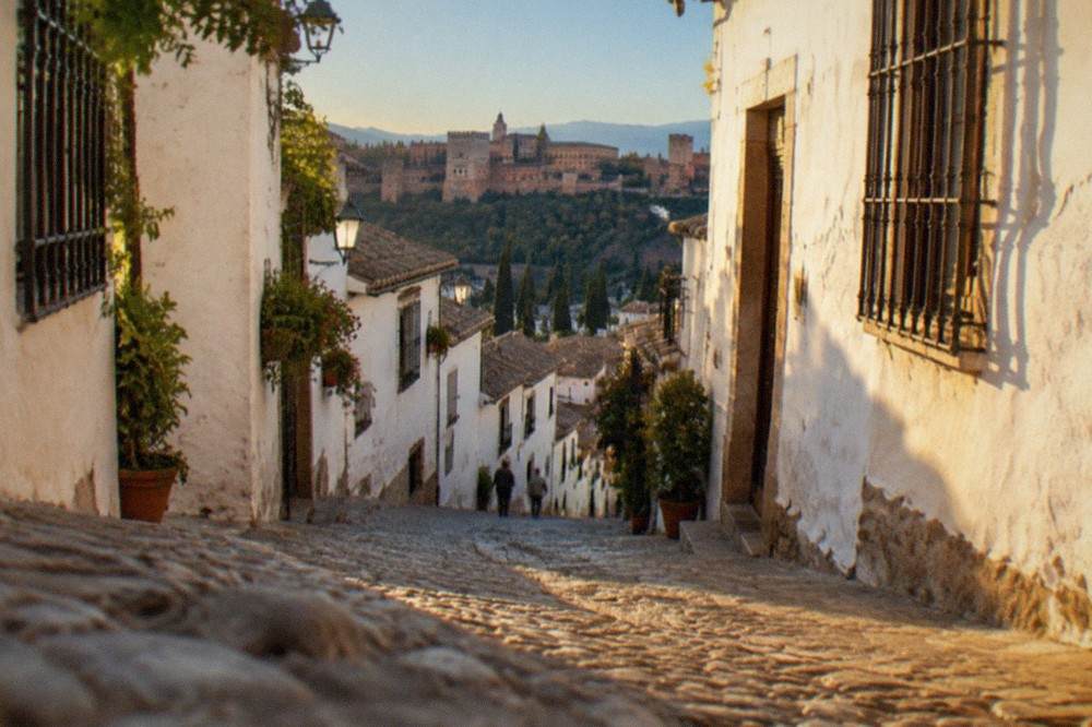 A narrow street in the Albaicin quarter of Granada at sunset with the Alhambra visible across the valley