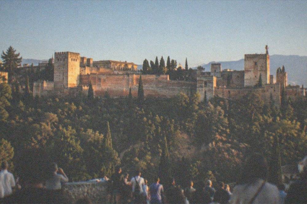 Crowd gathered at a plaza viewpoint at dusk with the Alhambra lit in warm light