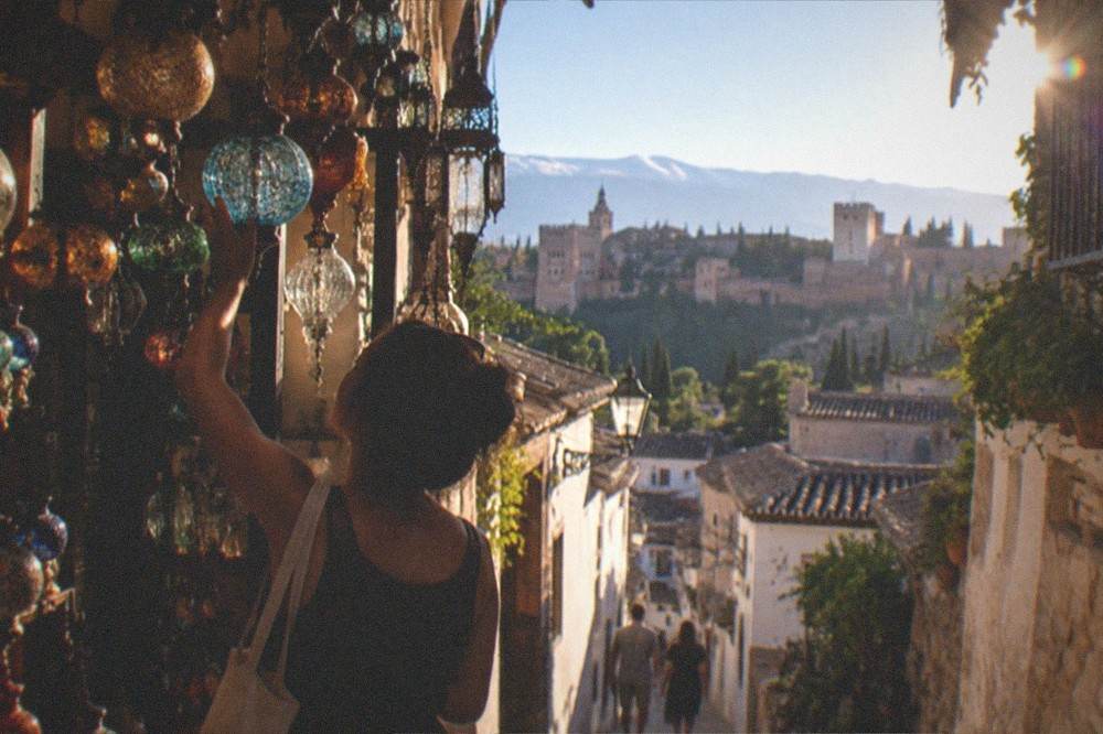 A shopper examining a hanging glass lamp inside a narrow Granada bazaar arcade