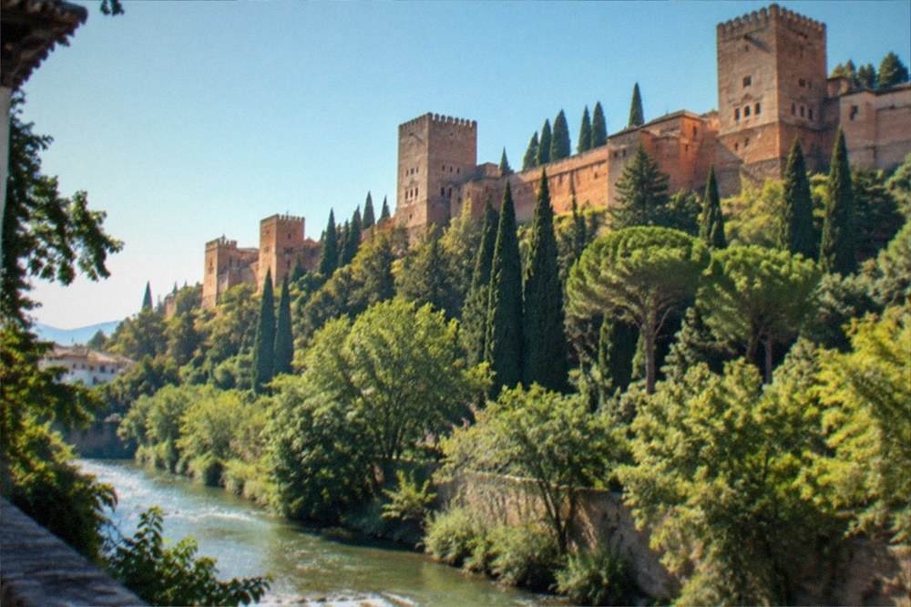 The Alhambra fortress walls seen from the Darro valley with trees along the river below