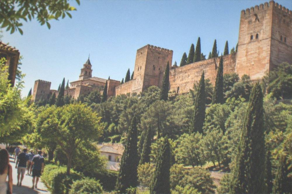 Visitors walking along a tree-lined path inside the Alhambra grounds with a palace tower behind