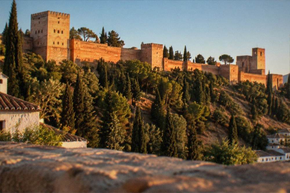 The Alhambra walls glowing red at sunset seen from a hillside across the valley