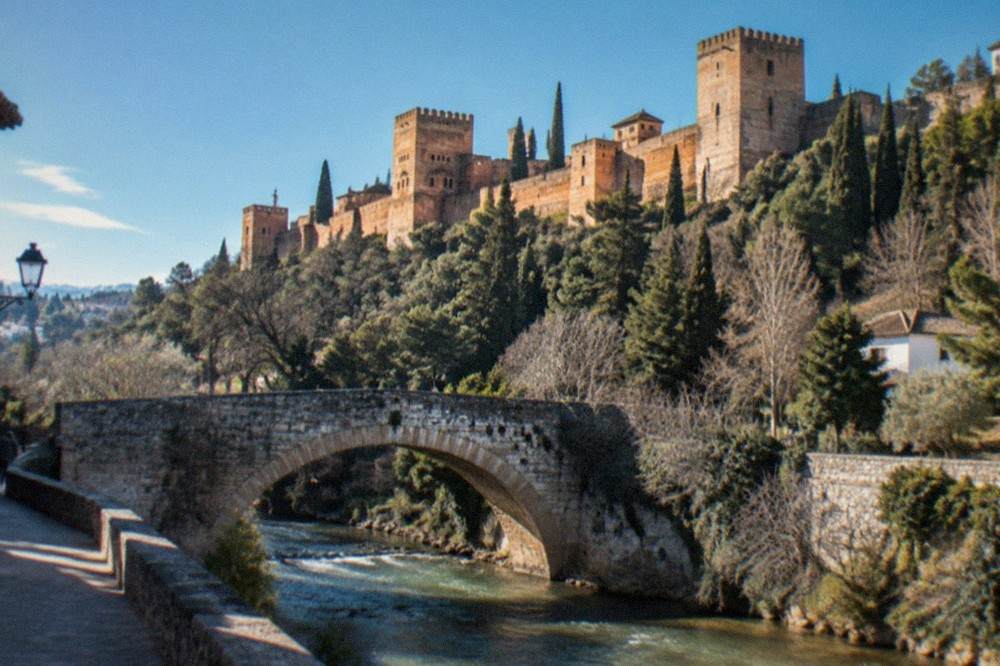 An old stone bridge over the Darro river with the Alhambra walls visible above the trees