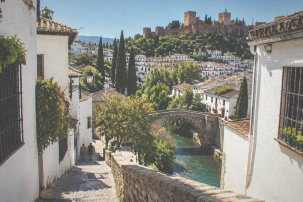 A narrow stone bridge over the Darro river with white houses of the Albaicin on the hillside above