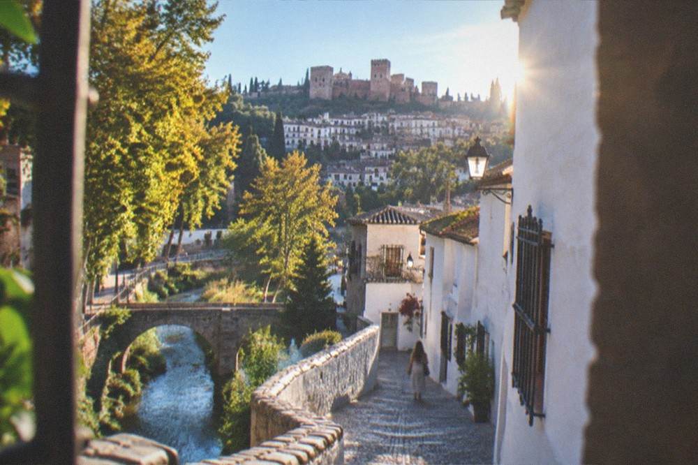 A view along the river walk with stone bridges, plane trees and the hillside houses of the Albaicin