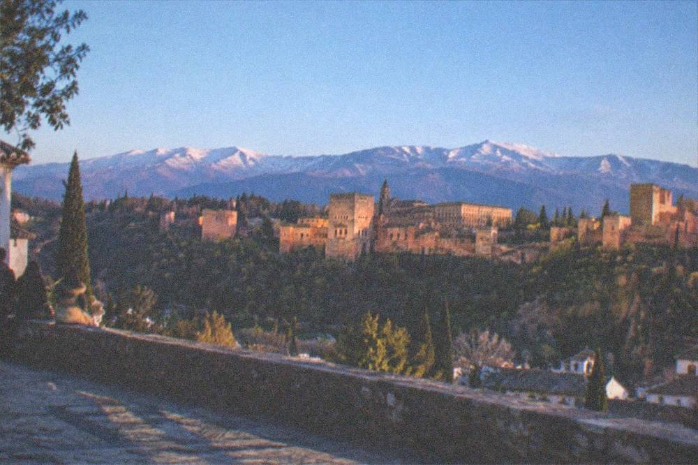 Panorama of a reddish hilltop palace complex with gardens and a mountain range behind it
