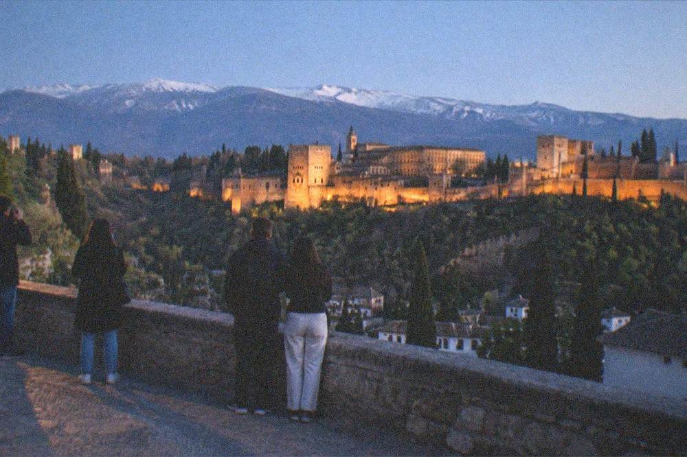 People with cameras and phones at a stone railing at dusk with a lit palace across a valley