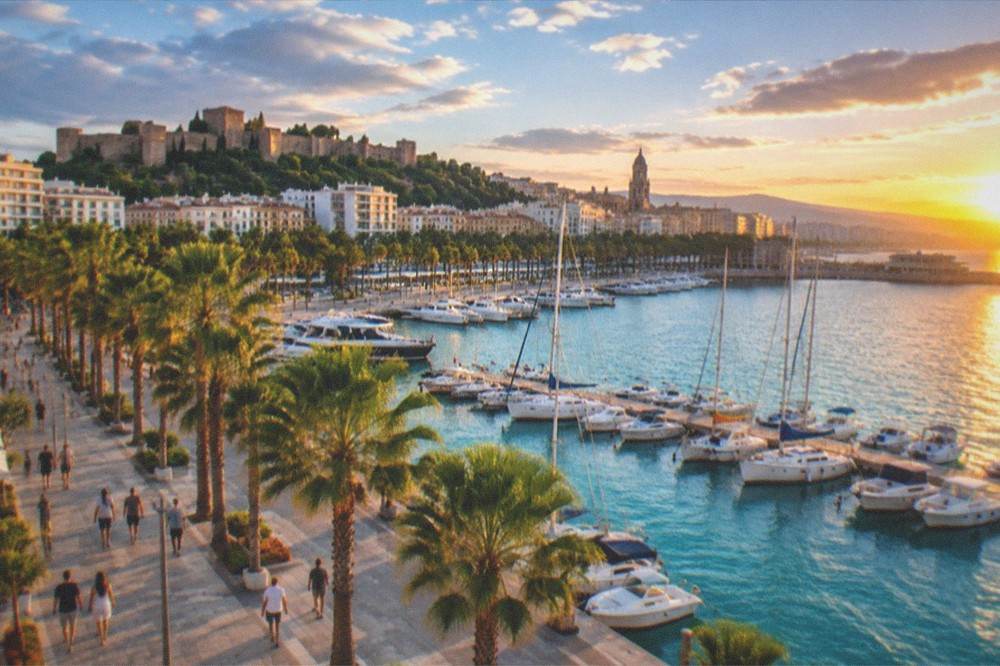 Sunset over a Mediterranean coastal city with palm-lined promenade and harbour lights