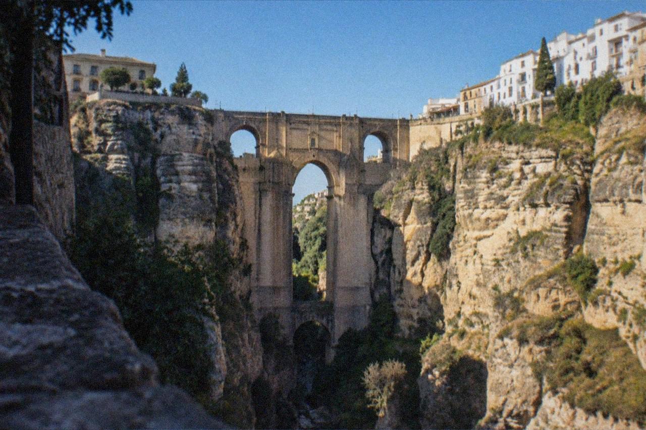 The stone arches of a bridge spanning a deep rocky gorge with white buildings on both cliff edges under a clear sky