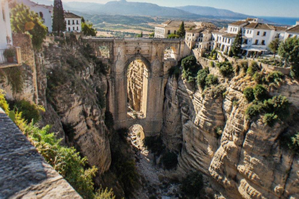 A narrow stone bridge arching over a deep gorge with a dry riverbed far below and cliffs rising on both sides