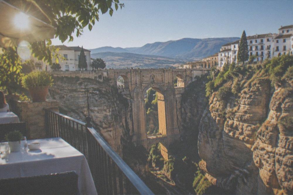 A shaded restaurant terrace with white tablecloths overlooking a deep gorge and distant hills