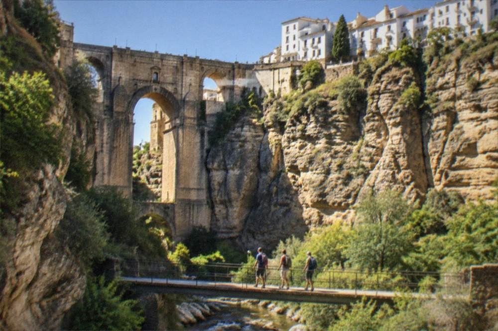 Hikers crossing a narrow footbridge over a mountain stream in a green shaded gorge