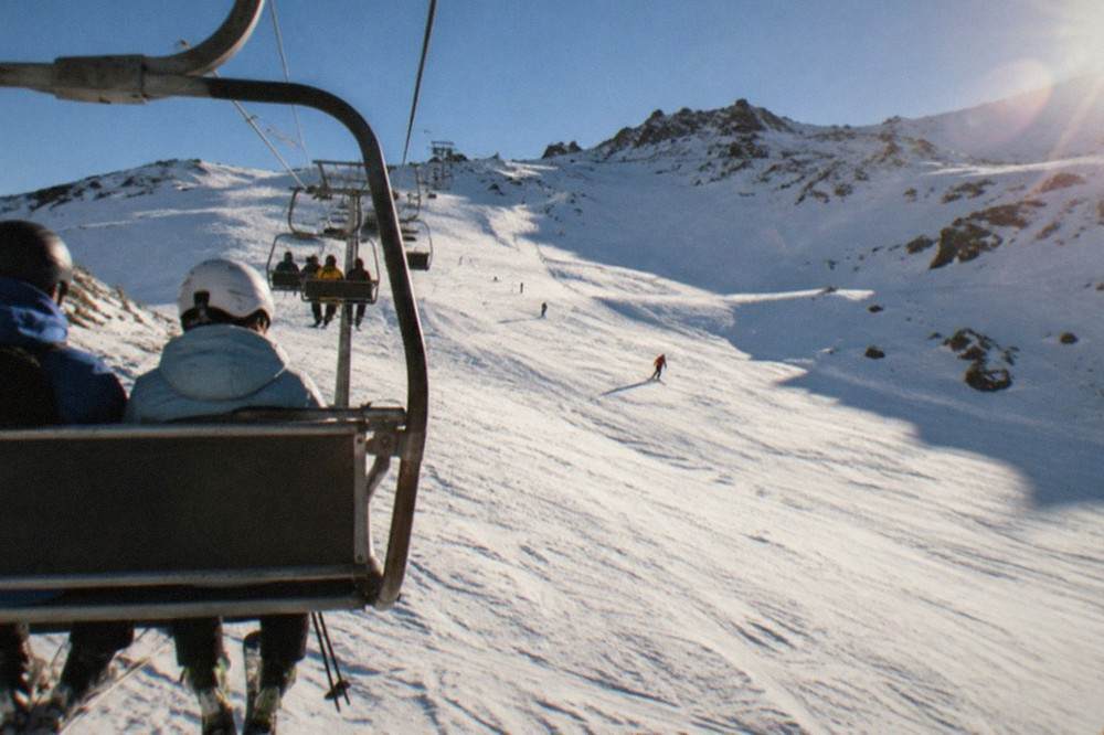 Skiers riding a chairlift up a wide white piste on a sunny morning