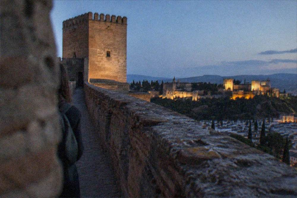 The floodlit towers of the Alcazaba seen from a dark garden path below