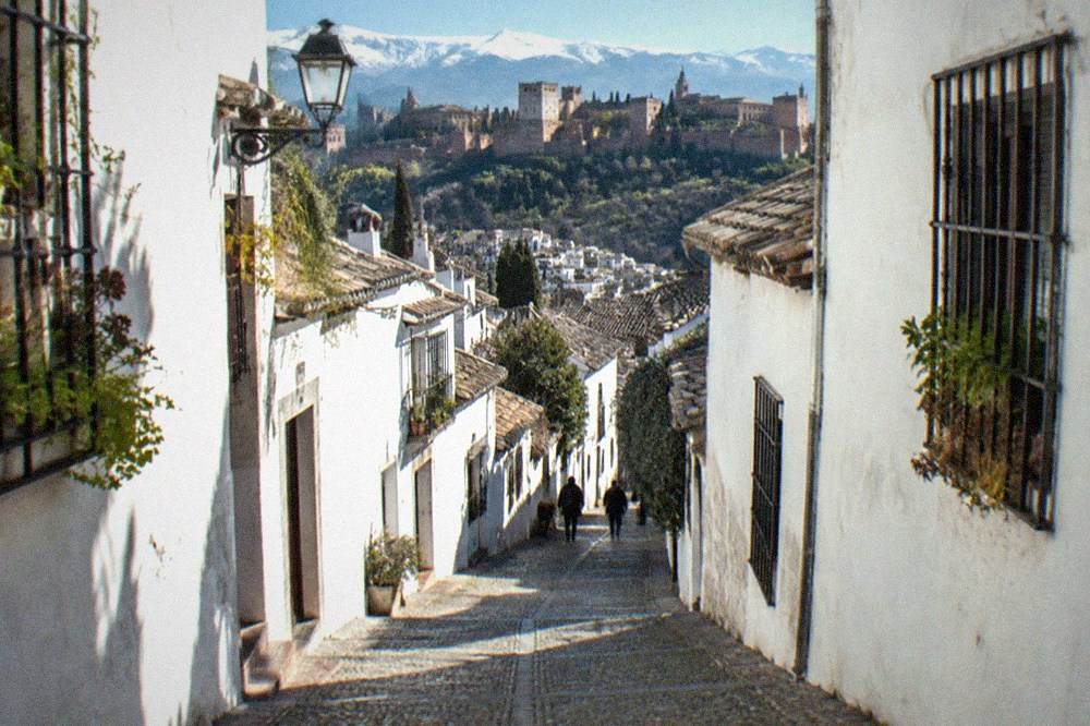 A narrow cobbled street in the Albaicin district with whitewashed houses and the Sierra Nevada visible in the distance