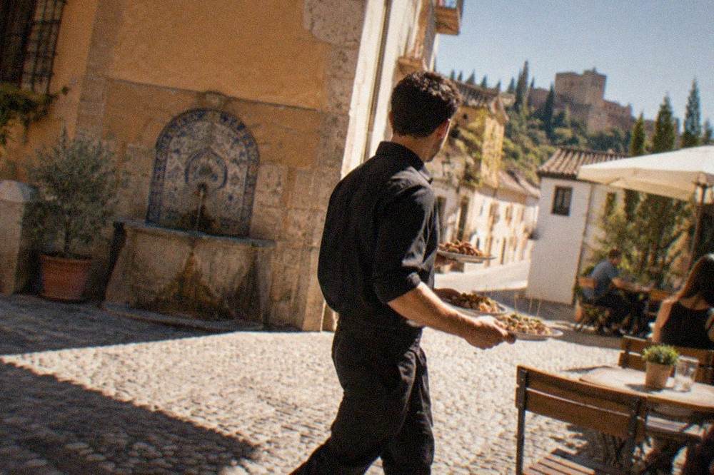 A waiter carrying plates of tapas to a table on a sunlit square in the Realejo district