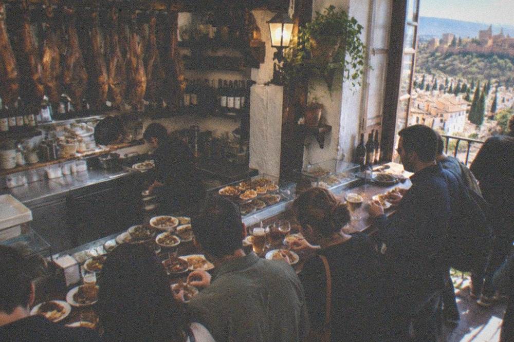 Locals standing at a crowded bar counter eating tapas in a traditional Granada tavern
