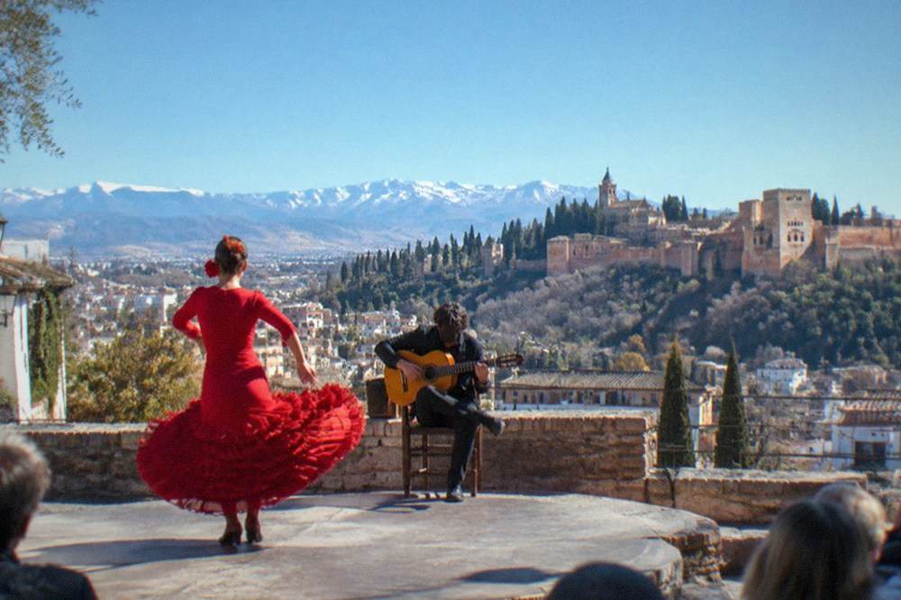 A dancer in a red dress mid-turn on a small stone stage with a guitarist seated behind