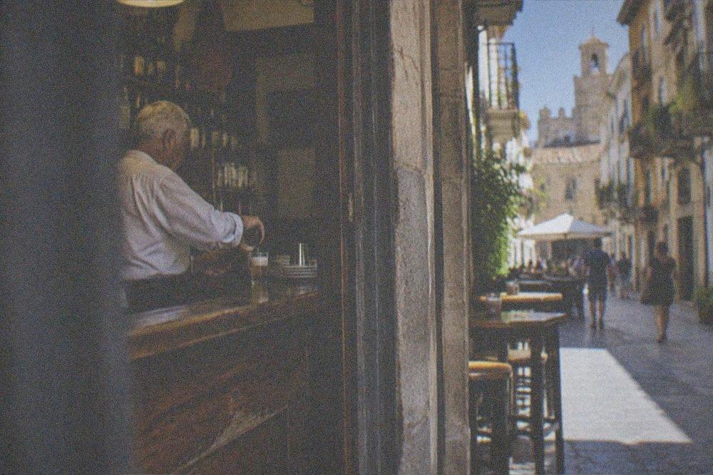 An elderly barman pouring a small beer behind a worn wooden counter in an old Granada tavern