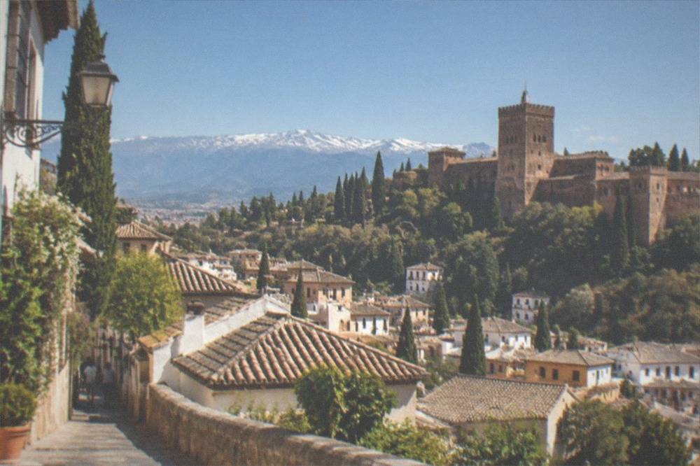 A red fortress tower rising above walled gardens with the roofs of a Spanish city below