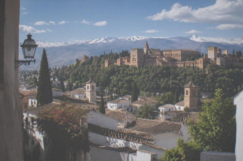 A view over whitewashed hillside houses and church towers toward a walled palace complex on a wooded ridge