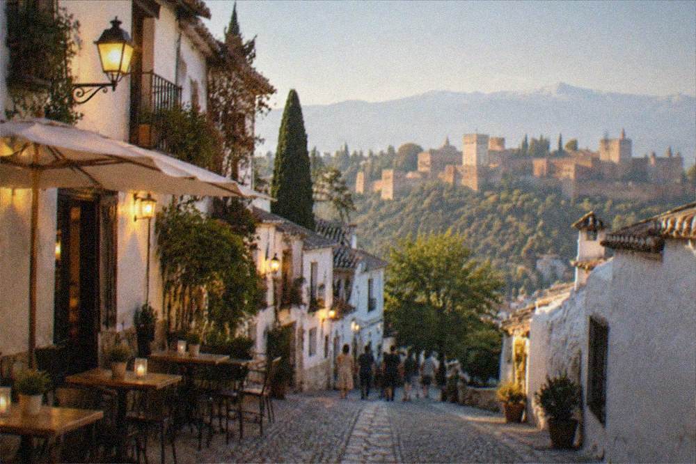 A narrow evening street of whitewashed buildings with warm lamplight and tables outside a small bar