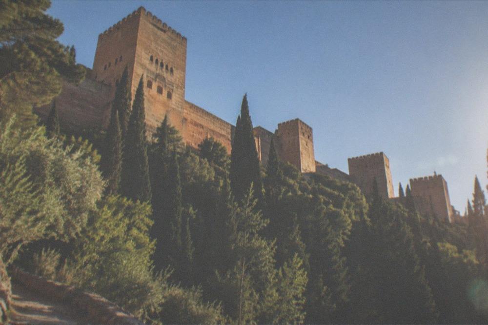 The red walls and towers of the Alhambra seen from a hillside path with cypress trees