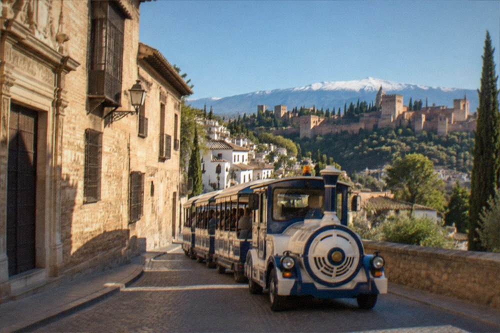 A tourist train passing a historic stone building on a narrow Granada street