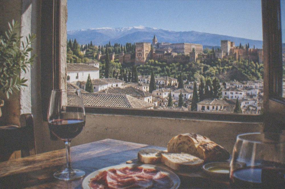 A table of cured ham, bread and red wine beside a window with a view over Granada rooftops