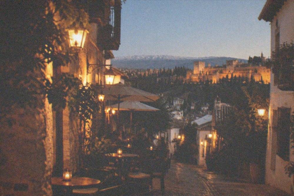 A narrow Granada street at night with small bar terraces lit by warm lamps