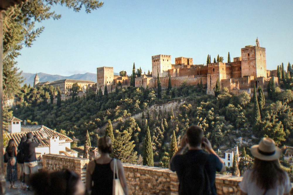 A crowded viewpoint terrace at sunset with visitors photographing the Alhambra across the valley
