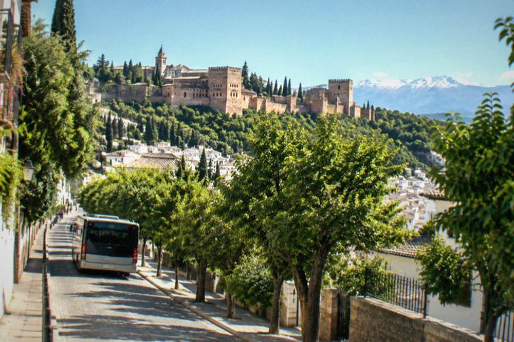 A city bus climbing a tree-lined street toward a hilltop fortress complex above Granada