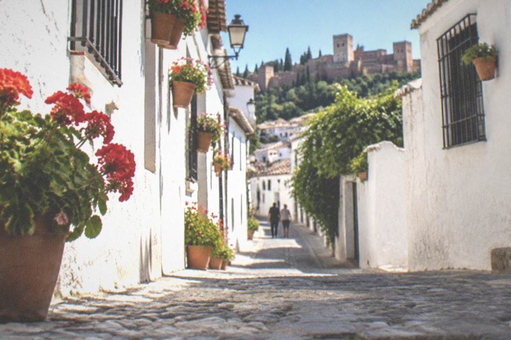 A narrow whitewashed street in the Albaicin quarter with potted geraniums and a view of hills beyond