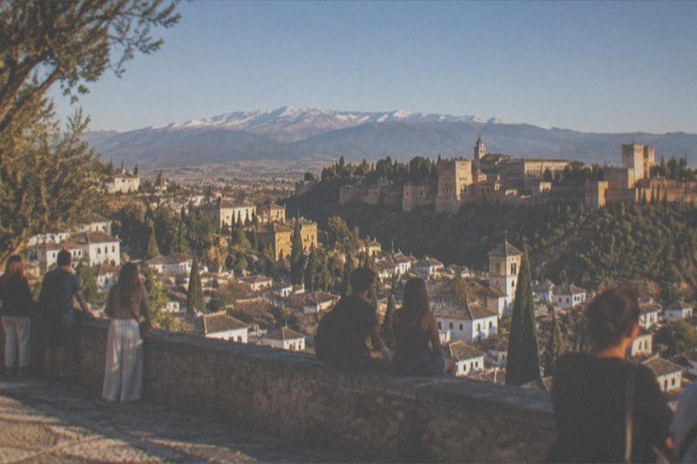 Visitors gathered at a hillside viewpoint watching the sunset over a Spanish city with a palace on the far hill