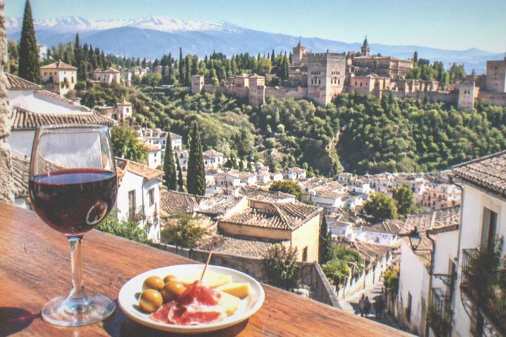 A small plate of tapas beside a glass of wine on a wooden bar counter in Granada