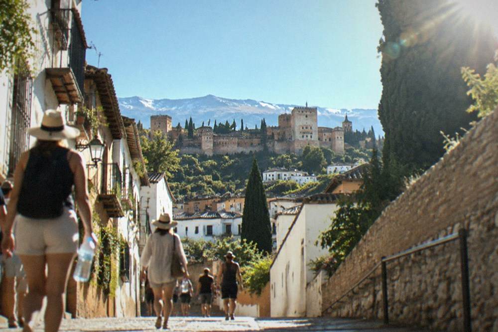 People walking with sun hats and water bottles along a sunlit Granada street in high summer