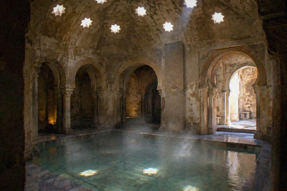 Sunken stone pool with steaming water under star-shaped ceiling skylights in a dim bath house