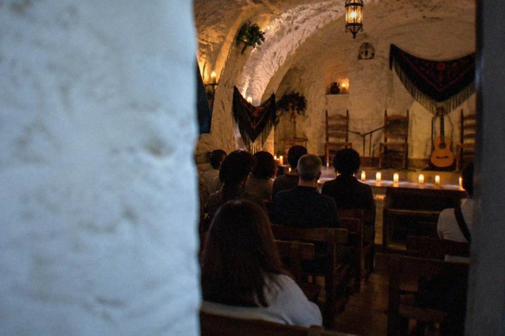 A small audience seated on wooden benches in a candle-lit cave chamber waiting for a flamenco performance to begin