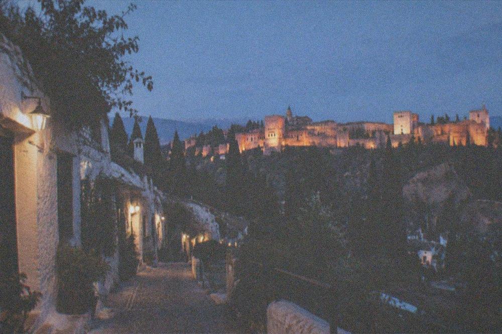 A narrow lane in Sacromonte at night, cave-house doors lit by small lamps with the Alhambra illuminated across the valley